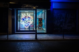 A tattoo parlor with a brightly lit neon sign in the window advertising 'Cesar Tattoos Lopez' in a heart shape with a crown on top. The shop interior is visible through the window, showing a chair and a colorful piece of artwork on a stand. The exterior of the shop is dark, with blue lighting creating a contrasting atmosphere.