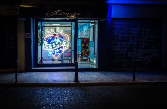 A tattoo parlor with a brightly lit neon sign in the window advertising 'Cesar Tattoos Lopez' in a heart shape with a crown on top. The shop interior is visible through the window, showing a chair and a colorful piece of artwork on a stand. The exterior of the shop is dark, with blue lighting creating a contrasting atmosphere.