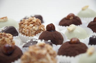 An assortment of gourmet truffles arranged on a white background. Each truffle is uniquely decorated, some with nuts, others with cocoa powder, and some with white chocolate coatings. They are placed in paper wrappers, enhancing their presentation.