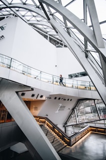 A modern architectural interior with sleek lines and an industrial design. An angular staircase with illuminated handrails leads down to a glass-walled area, which provides an outside view. A person is seen walking along a glass railing. The structural supports and ceiling feature intricate metal beams.