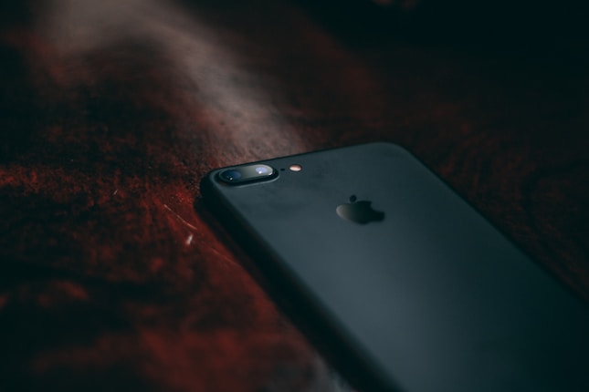A black smartphone lies on a dark wooden surface. The low lighting casts shadows and highlights the sleek, matte finish of the device. A small camera and flash are visible on the upper portion of the back.