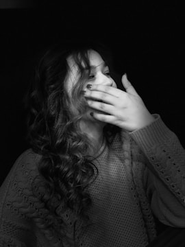 A woman with curly hair is captured in grayscale, wearing a textured sweater. Her hand partially covers her face, which appears to be expressing amusement or surprise.