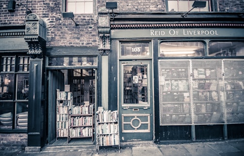 The image depicts the exterior of a vintage bookstore with a dark, slightly weathered facade. A collection of books is displayed outside on wooden racks beside the entrance. The sign above reads 'Reid of Liverpool.' The building features intricate architectural details with a brick exterior and tall windows.