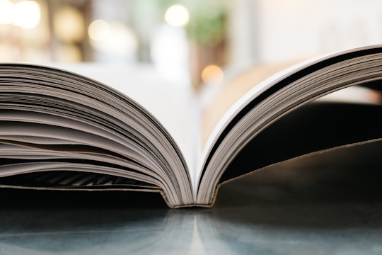 A close-up view of an open book with a focus on the curved pages resting on a flat surface. The background is softly blurred, providing a warm and inviting atmosphere.