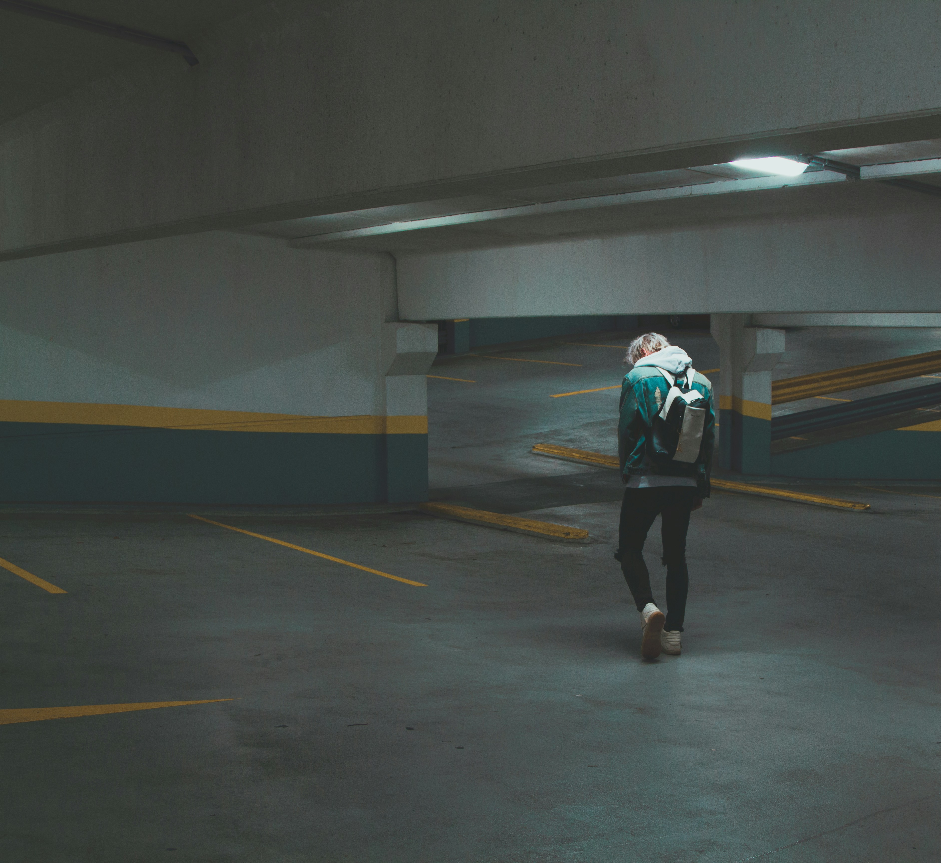 back of person in jacket over hoodie, jeans, sneakers, and a backpack walking in an empty parking garage