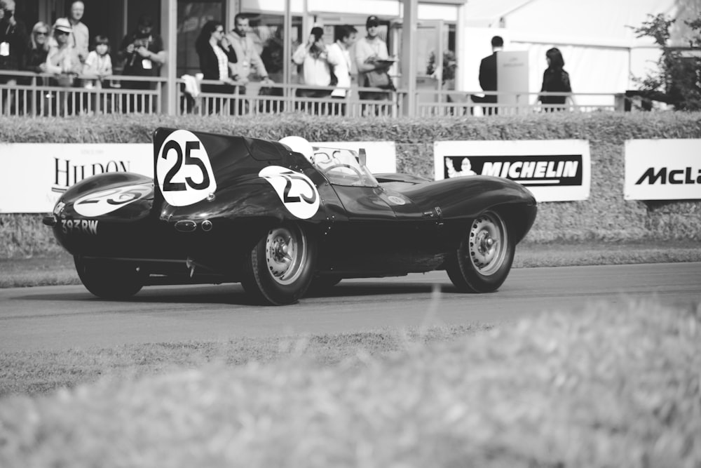 grayscale photo of classic stock car on road near people standing beside rails