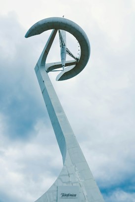A modernist tower structure with a spiral design against a backdrop of cloudy skies. The structure is predominantly white, featuring a sleek, curving form that extends upward. A label at the base reads 'Telef&oacute;nica'. The cloudy sky adds a sense of depth and contrast to the architectural design.