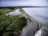 Aerial view of a coastal landscape featuring a long, narrow strip of beach separating the ocean from a series of lush green fields and wetlands. The image shows the meeting point of the land and sea, with scattered logs visible on the sandy beach. The water is a calm, muted blue, while patches of white clouds are visible in the sky.