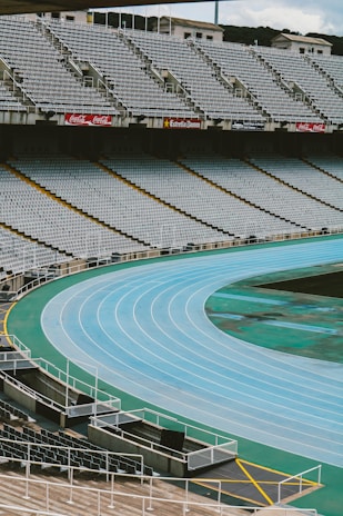 An empty stadium with a blue running track encircling a green field. The stands are filled with rows of empty gray seats, and there are billboards with advertisements above the top row. The overall environment appears calm and orderly, ready for an event but currently unoccupied.