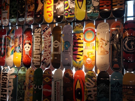 A collection of skateboard decks is displayed, each featuring unique and vibrant designs. The decks have various graphics ranging from artistic illustrations and logos to characters and colorful patterns. They're arranged vertically against a lighted backdrop, highlighting the diversity in style and creativity.