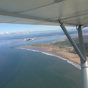 A small airplane is flying above a coastline with an expansive view of the ocean and shoreline. The wing and struts of a second aircraft are visible in the top and right edges of the image, indicating the photo was taken from another plane. The coastline features sandy beaches and some green vegetation, with gentle waves approaching the shore. The sky is mostly clear with a few scattered clouds.