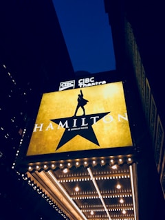 A theater marquee displays a large illuminated sign for the musical 'Hamilton' with a silhouette of a person raising their hand inside a star. The background is dark, and the marquee lights are glowing brightly against the night sky.