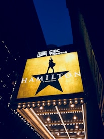 A theater marquee displays a large illuminated sign for the musical 'Hamilton' with a silhouette of a person raising their hand inside a star. The background is dark, and the marquee lights are glowing brightly against the night sky.