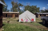 A white tent is set up on grassy ground with red and pink chairs in front of it. Surrounding the tent are rustic wooden cabins with weathered exteriors under a blue sky. A dog is walking nearby, and a black truck is parked to the right of the tent.