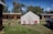 A white tent is set up on grassy ground with red and pink chairs in front of it. Surrounding the tent are rustic wooden cabins with weathered exteriors under a blue sky. A dog is walking nearby, and a black truck is parked to the right of the tent.
