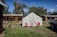 A white tent is set up on grassy ground with red and pink chairs in front of it. Surrounding the tent are rustic wooden cabins with weathered exteriors under a blue sky. A dog is walking nearby, and a black truck is parked to the right of the tent.