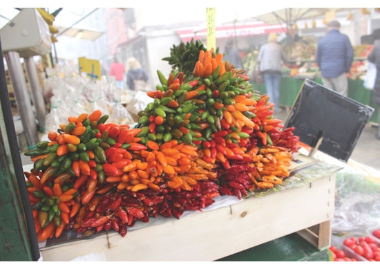 A vibrant display of assorted peppers in shades of red, orange, and green is arranged on a wooden stand at an outdoor market. People and other market stalls are visible in the slightly blurred background, indicating a bustling market atmosphere.