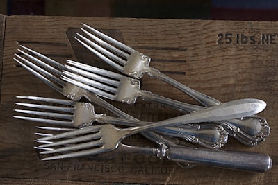 A collection of silver forks and knives with ornate handles are placed on a wooden surface. The wood shows signs of wear and contains text reading '25 lbs. NE' along with 'San Francisco, California'. The forks are arranged in an overlapping pattern, creating a sense of casual disarray.