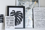 A collection of framed artwork and books on a shelf. The central piece is a black and white framed print of a monstera leaf. To the left, a book titled 'WHATEVER YOU THINK THINK THE OPPOSITE' by Paul Arden is propped up in front of the artwork. On the right, there's a partially visible book or magazine with handwritten text and headings in a creative, artistic style. The background includes abstract art with hints of gold and muted colors.