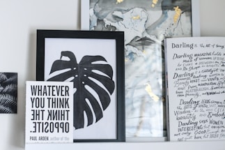 A collection of framed artwork and books on a shelf. The central piece is a black and white framed print of a monstera leaf. To the left, a book titled 'WHATEVER YOU THINK THINK THE OPPOSITE' by Paul Arden is propped up in front of the artwork. On the right, there's a partially visible book or magazine with handwritten text and headings in a creative, artistic style. The background includes abstract art with hints of gold and muted colors.