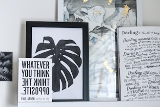 A collection of framed artwork and books on a shelf. The central piece is a black and white framed print of a monstera leaf. To the left, a book titled 'WHATEVER YOU THINK THINK THE OPPOSITE' by Paul Arden is propped up in front of the artwork. On the right, there's a partially visible book or magazine with handwritten text and headings in a creative, artistic style. The background includes abstract art with hints of gold and muted colors.