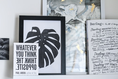 A collection of framed artwork and books on a shelf. The central piece is a black and white framed print of a monstera leaf. To the left, a book titled 'WHATEVER YOU THINK THINK THE OPPOSITE' by Paul Arden is propped up in front of the artwork. On the right, there's a partially visible book or magazine with handwritten text and headings in a creative, artistic style. The background includes abstract art with hints of gold and muted colors.
