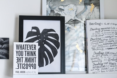 A collection of framed artwork and books on a shelf. The central piece is a black and white framed print of a monstera leaf. To the left, a book titled 'WHATEVER YOU THINK THINK THE OPPOSITE' by Paul Arden is propped up in front of the artwork. On the right, there's a partially visible book or magazine with handwritten text and headings in a creative, artistic style. The background includes abstract art with hints of gold and muted colors.