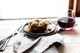 A plate with a cooked dish garnished with sautéed onions and vegetables is set on a soft cloth. Next to it is a glass of red wine. The setting suggests a cozy and rustic atmosphere.