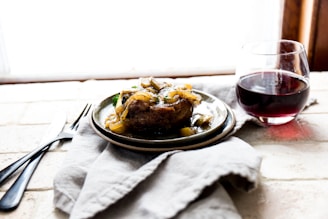 A plate with a cooked dish garnished with sautéed onions and vegetables is set on a soft cloth. Next to it is a glass of red wine. The setting suggests a cozy and rustic atmosphere.