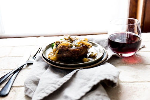 A plate with a cooked dish garnished with saut&eacute;ed onions and vegetables is set on a soft cloth. Next to it is a glass of red wine. The setting suggests a cozy and rustic atmosphere.