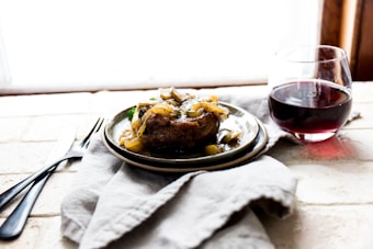 A plate with a cooked dish garnished with saut&eacute;ed onions and vegetables is set on a soft cloth. Next to it is a glass of red wine. The setting suggests a cozy and rustic atmosphere.