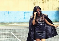 A woman is standing confidently by a tennis net, wearing sunglasses, a black jacket with leopard print on the shoulders, and a black shirt with the word 'PARIS'. The background features a blue and beige wall with some weathered textures.