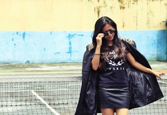 A woman is standing confidently by a tennis net, wearing sunglasses, a black jacket with leopard print on the shoulders, and a black shirt with the word 'PARIS'. The background features a blue and beige wall with some weathered textures.
