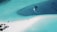 Aerial view of a turquoise lagoon with a sandy beach forming a narrow strip between the water. Two boats are moving along the clear water, leaving trails behind. On the left side, small thatched-roof structures and some greenery are visible.