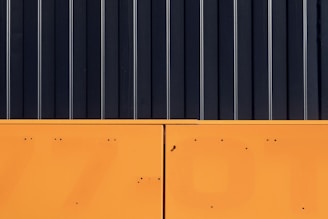 The image features a minimalist composition with a stark contrast between two major sections. The upper part consists of vertical black panels with thin white lines running through them, resembling a modern industrial design. The lower section is a solid block of bright orange with small dark spots and lines.