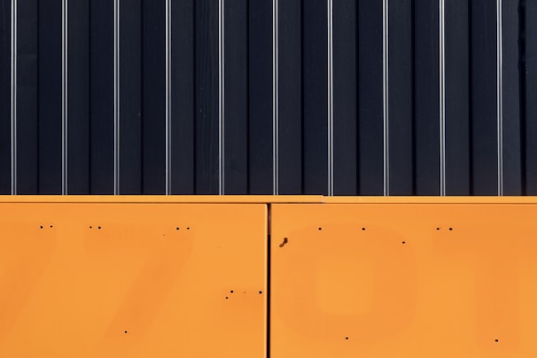 The image features a minimalist composition with a stark contrast between two major sections. The upper part consists of vertical black panels with thin white lines running through them, resembling a modern industrial design. The lower section is a solid block of bright orange with small dark spots and lines.
