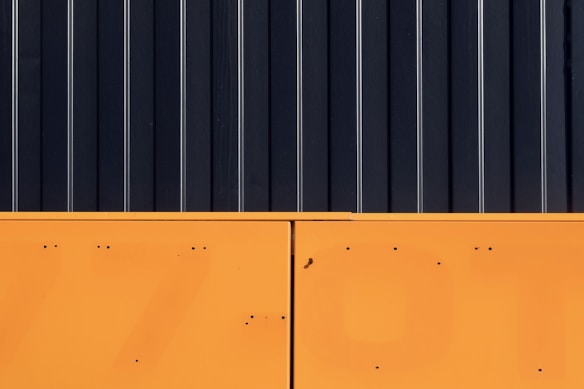 The image features a minimalist composition with a stark contrast between two major sections. The upper part consists of vertical black panels with thin white lines running through them, resembling a modern industrial design. The lower section is a solid block of bright orange with small dark spots and lines.