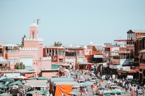 A bustling street scene in a Middle Eastern or North African town, characterized by traditional buildings with pinkish-red facades. The street is crowded with people and vehicles, including cars and motorbikes. Market stalls with green and red canopy tarps line the streets, with goods on display. Satellite dishes are visible on the rooftops, and the background features a mosque with a tower and intricate architecture.