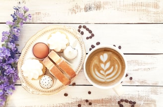 A latte with artful foam in a white cup sits on a rustic wooden surface, paired with an ornate plate featuring pastries, biscotti, a boiled egg, and some whipped cream dollops. Purple flowers and scattered coffee beans add to the aesthetic presentation.