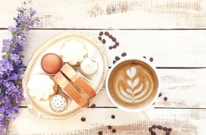 A latte with artful foam in a white cup sits on a rustic wooden surface, paired with an ornate plate featuring pastries, biscotti, a boiled egg, and some whipped cream dollops. Purple flowers and scattered coffee beans add to the aesthetic presentation.