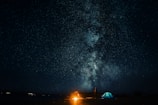 A starry night sky filled with a bright, detailed view of the Milky Way galaxy. Below, a campfire illuminates the darkness, with a tent and a person standing nearby. The scene conveys a sense of peaceful solitude and wonder.