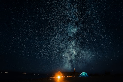 A starry night sky filled with a bright, detailed view of the Milky Way galaxy. Below, a campfire illuminates the darkness, with a tent and a person standing nearby. The scene conveys a sense of peaceful solitude and wonder.