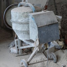 A cement mixer covered in dust, situated in a construction site. Surrounding the mixer are bags of cement, a shovel, and various construction materials scattered around.