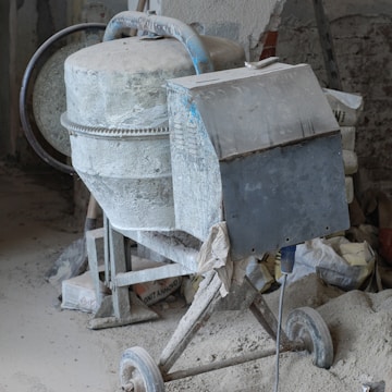 A cement mixer covered in dust, situated in a construction site. Surrounding the mixer are bags of cement, a shovel, and various construction materials scattered around.
