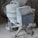 A cement mixer covered in dust, situated in a construction site. Surrounding the mixer are bags of cement, a shovel, and various construction materials scattered around.