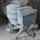 A cement mixer covered in dust, situated in a construction site. Surrounding the mixer are bags of cement, a shovel, and various construction materials scattered around.