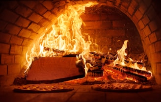 A brick oven with a roaring fire inside, surrounded by intense flames and glowing embers. Positioned at the base of the oven are a couple of pizzas being cooked amid the heat.