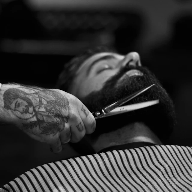Black and white photo of a person trimming a man's beard with scissors