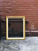 A wooden picture frame with a gold finish leans against a textured red brick wall. The frame is empty and reflects the surrounding environment, including some pipes and a shadow. The ground is made of worn concrete, displaying some stains and marks.