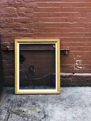 A wooden picture frame with a gold finish leans against a textured red brick wall. The frame is empty and reflects the surrounding environment, including some pipes and a shadow. The ground is made of worn concrete, displaying some stains and marks.